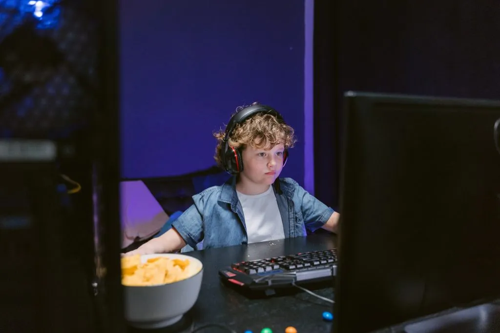 Um menino se senta em uma mesa atrás de monitores de computador jogando jogo de computador