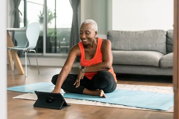 Senior woman assis sur un tapis de yoga dans un salon, regardant un ordinateur tablette