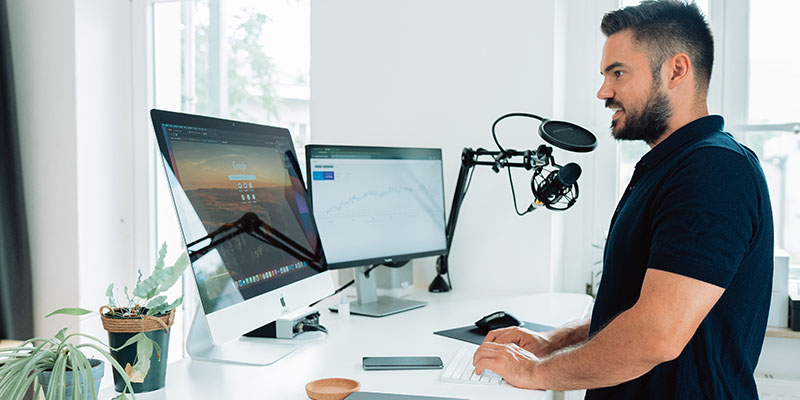 Bir sosyal medya ajansında masanın önünde podcast mikrofonu ve masasında bölünmüş ekranlı bilgisayar ve dizüstü bilgisayar bulunan iki ekranla çalışan bir adam.