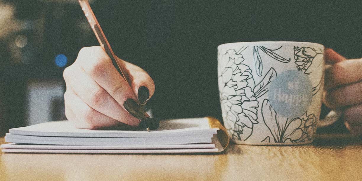 Persona escribiendo en un cuaderno con una taza de café sobre la mesa