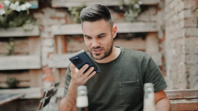 Ein Mann schaut auf sein Telefon und spricht über den Lautsprecher, während er an einem Tisch sitzt.